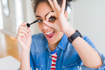 Young african american girl applying eyelashes mascara with happy face smiling doing ok sign with hand on eye looking through fingers