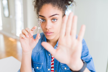 Young african american girl using eyelashes curler with open hand doing stop sign with serious and confident expression, defense gesture