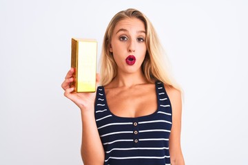 Young beautiful blond woman holding gold ingot standing over isolated white background scared in shock with a surprise face, afraid and excited with fear expression