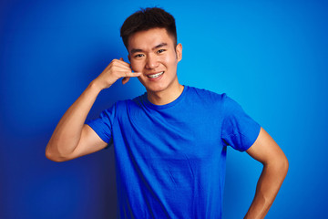 Young asian chinese man wearing t-shirt standing over isolated blue background smiling doing phone gesture with hand and fingers like talking on the telephone. Communicating concepts.