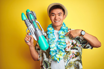 Asian chinese man wearing floral hawaian lei and water gun over isolated yellow background with...