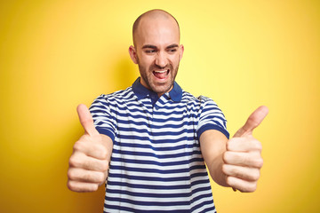 Young bald man with beard wearing casual striped blue t-shirt over yellow isolated background approving doing positive gesture with hand, thumbs up smiling and happy for success. Winner gesture.