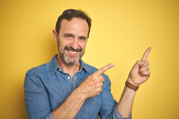 Handsome middle age senior man with grey hair over isolated yellow background smiling and looking at the camera pointing with two hands and fingers to the side.