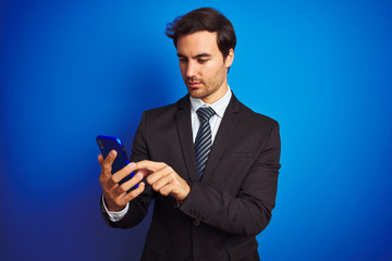 Young handsome businessman using smartphone standing over isolated blue background with a confident expression on smart face thinking serious