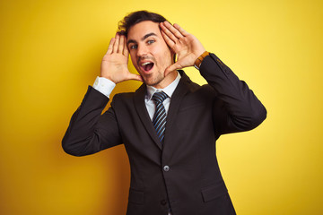 Young handsome businessman wearing suit and tie standing over isolated yellow background Smiling cheerful playing peek a boo with hands showing face. Surprised and exited