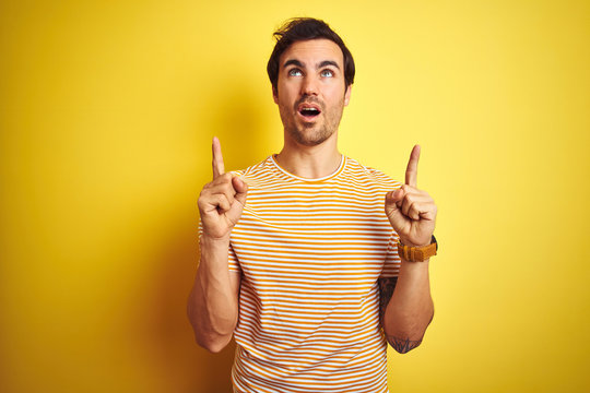 Young handsome man with tattoo wearing striped t-shirt over isolated yellow background amazed and surprised looking up and pointing with fingers and raised arms.