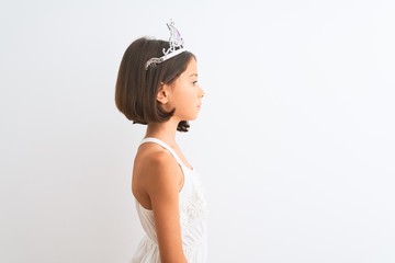 Beautiful child girl wearing princess crown standing over isolated white background looking to...