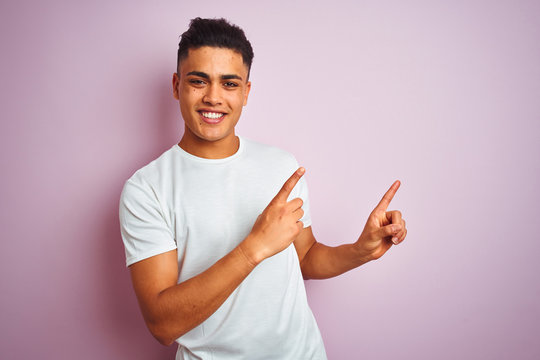 Young brazilian man wearing t-shirt standing over isolated pink background smiling and looking at the camera pointing with two hands and fingers to the side.