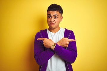 Young brazilian man wearing purple sweatshirt standing over isolated yellow background Pointing to both sides with fingers, different direction disagree