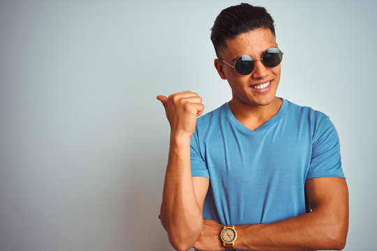 Young brazilian man wearing blue t-shirt and sunglasses over isolated white background smiling with happy face looking and pointing to the side with thumb up.