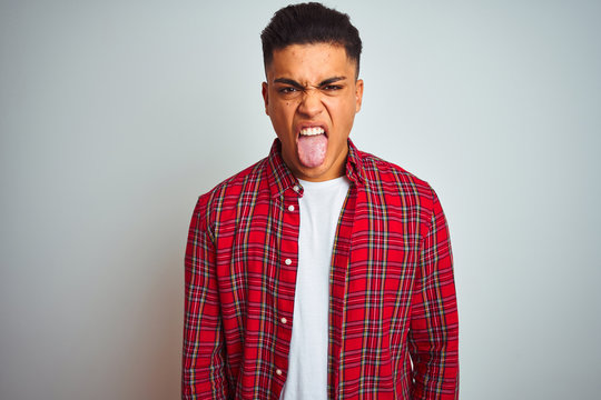 Young brazilian man wearing red shirt standing over isolated white background sticking tongue out happy with funny expression. Emotion concept.