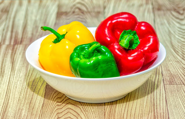 Red, green & yellow bell peppers (capsicum) in a white bowl on a wooden table.