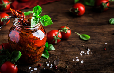 Italian Sun Dried tomatoes in olive oil with green basil and spices in glass jar on wooden kitchen table, rustic style, copy space