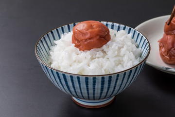 黒背景に白いお米と梅干しと後ろにお皿にのっている梅干し