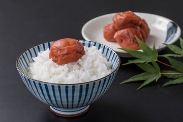 黒背景に白いお米と梅干しとバックにお皿にのっている梅干しと葉っぱ緑