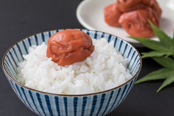 黒背景に白いお米と梅干しとバックにお皿にのっている梅干しと紅葉の葉っぱ緑