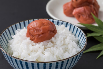 黒背景に白いお米と梅干しとバックにお皿にのっている梅干しと紅葉の葉っぱ緑