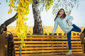 Sports girl in light down jacket doing stretching legs on bench on morning of autumn.