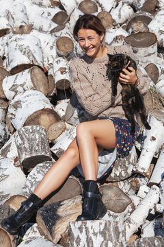 woman in a knitted hat, knitting sweater, pretty dress and boots, smiles cute, holding a black cat and sits on birch logs. The concept of rustic style and life in ancient times