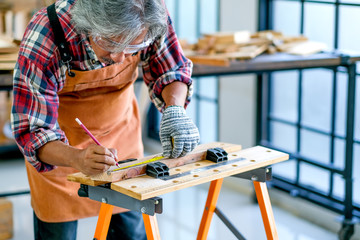 Elder Asian craftsman act as measurement of wood in the room with wood pile and surround with glasses.