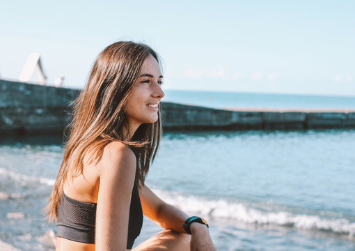Young slim athletic long hair smiling woman in sportswear sitting on the sea beach, healthy lifestyle
