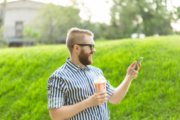 Naklejka na ściany i meble Side view of a stylish young man with glasses and a beard with a milkshake in his hands talking on video with his friends using smartphone and high-speed free Internet in the park on a warm summer day
