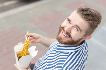 Young male hipster with a mustache and beard is eating chinese noodles with wooden chopsticks from a white lunch box. Asian cuisine concept.