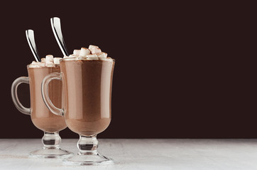 Elegant hot chocolate in two glass with marshmallows and silver spoon on dark brown background, copy space.