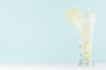 Frosty transparent lemonade with lemon slices, ice cubes, mineral water, yellow striped straw in elegant glass on white wood table, mint color wall.