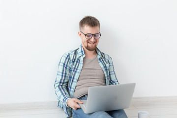 Positive young man in casual clothes surfing the Internet in search of new housing sitting on the floor in an empty room. The concept of finding an apartment using the Internet and a laptop.