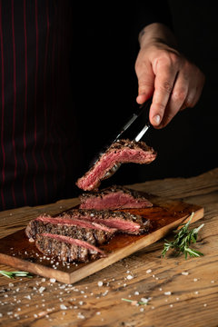 Slicing juicy beef steak by knife in chef hands closeup. Food cooking concept. Dark black background copy space.