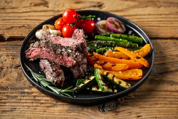 Poster Grilled and sliced beef steak with grilled vegetables served on black plate on old rustic wood background. © Anton