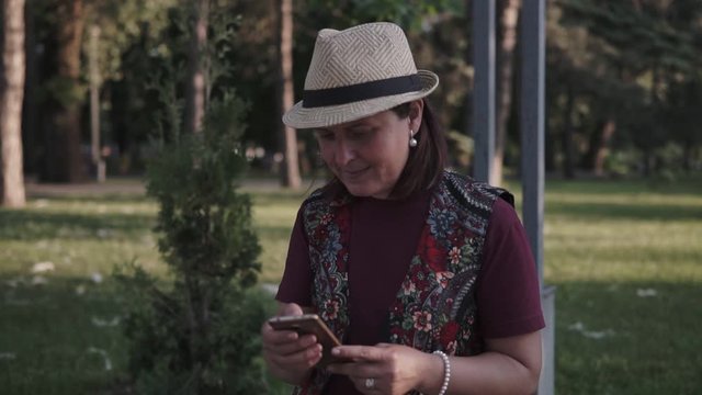 young brunette in a hat is using a mobile phone in a city summer park