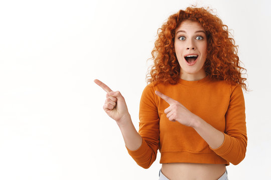 Cheerful amused attractive redhead woman with blue eyes, curly hair pointing left blank space, checking out cool promo, open mouth fascinated, look overwhelmed breathtaking event going nearby