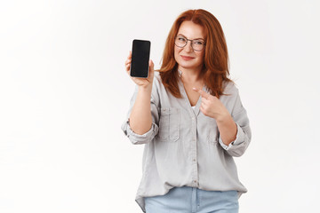 Fototapeta premium Better check out yourself. Assured successful middle-aged redhead businesswoman sharing professional secrets hold smartphone pointing phone screen promote app smiling delighted
