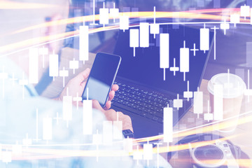 Candlestick chart in foreground. In background man uses smartphone, works on laptop. Businessman reads message in smartphone.