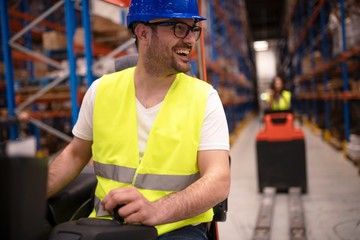Warehouse workers enjoy driving forklifts in large factory storage department. Industrial workers operating lifting machines to relocate palette with goods.