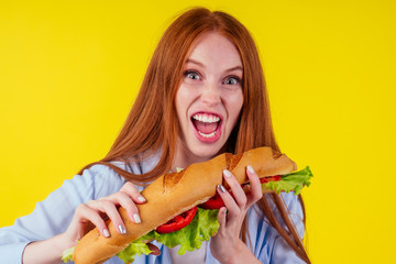Joyful funny redhaired ginger girl with wild eyes suffering from anarexia with greedy ,eating large sandwic in studio yellow background. eats after 6 pm