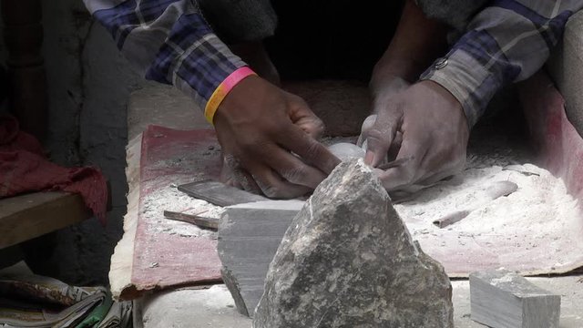 Young artist deftly carves a grey stone by tapping on his small chisel while securing it with his feet. Closeup.
