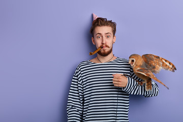 Handsome cheerful attarctive young man with owl in his arm having fun on party, blowing party horn...