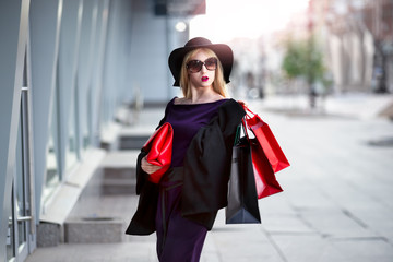 Fototapeta premium Stylish blonde woman in black coat, sunglasses and hat with shopping bags walks along the street