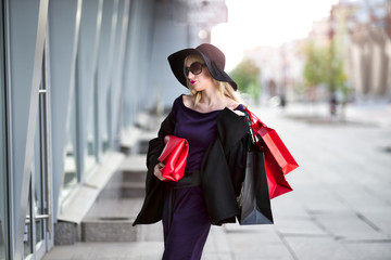 Fototapeta premium Stylish blonde woman in black coat, sunglasses and hat with shopping bags walks along the street