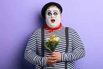 surprised shocked young man with painted face, striped clothes and suspenders with wide open mouth holding bouquet of artificial flowers.isolated blue background, studio shot