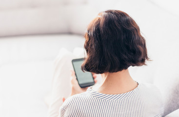 Middle-aged brunette woman with glasses on the gray sofa use smartphone.