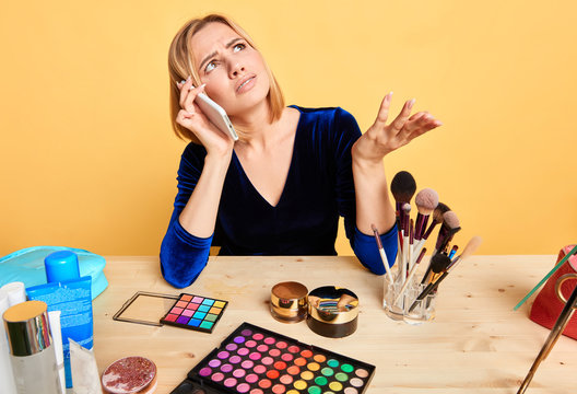 Portrait of dissatisfied young blonde female model talking by smartphone while making makeup and beauty routine, having unpleasant expression, frowning face, arguing and disagreeing, looking aside.