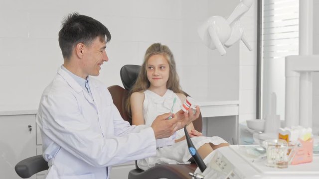 Cheerful male dentist showing little girl how to brush teeth. Charming little girl visiting dentist, learning brushing teeth properly. Young dentist working with child