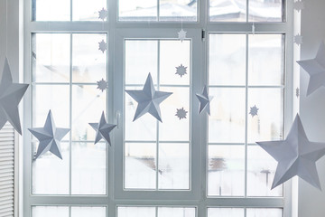 Large bright window. On the window-sill is a gray knitted plaid with gifts and sparkles with herlands. Paper Christmas stars are suspended on the background of the window.