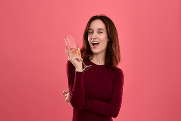 Excited pretty brunette girl holding wineglass of sparkling champagne standing isolated on a dark pink background and smiling at the camera.
