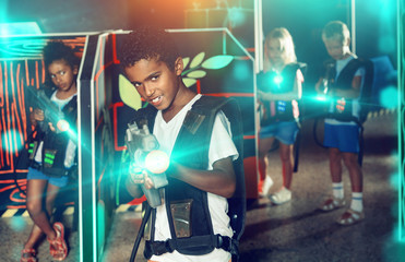 Emotional afro-american boy with laser pistol playing laser tag with friends on dark labyrinth