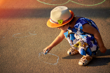 Cute baby girl in hat is drawing with chalk on asphalt in spring park.